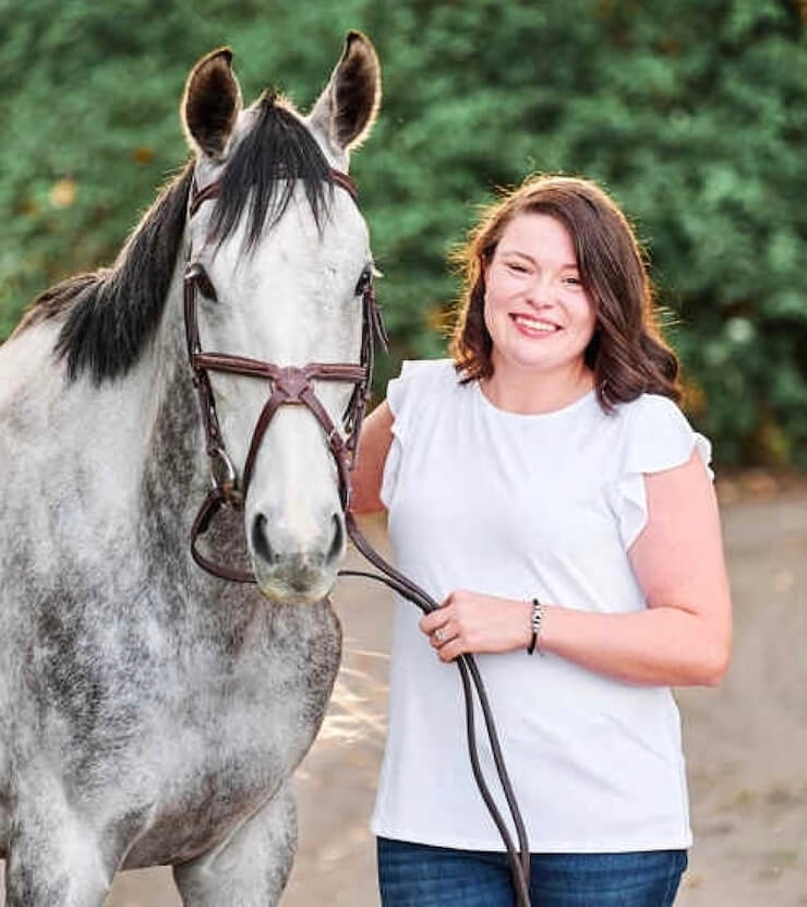 Katy McFarland, a brunette woman, smiling while standing next to a light grey and white horse.