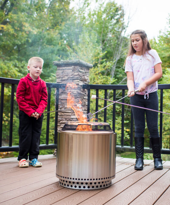 Two children standing outside next to a fireplace roasting marshmallows.